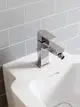 Chromfarbene Bidetarmatur von Ideal Standard mit ausladender Armatur und Wasserhahn. Die Montage erfolgt auf einem weißen Waschbecken vor grauen Fliesen.